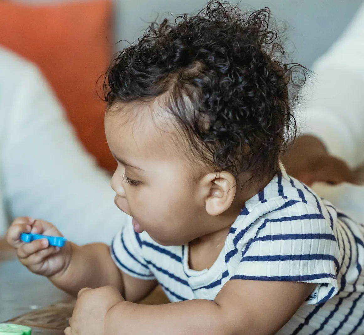 Curly-haired baby in a striped top leaning on a table, holding a small blue toy and exploring colourful play pieces